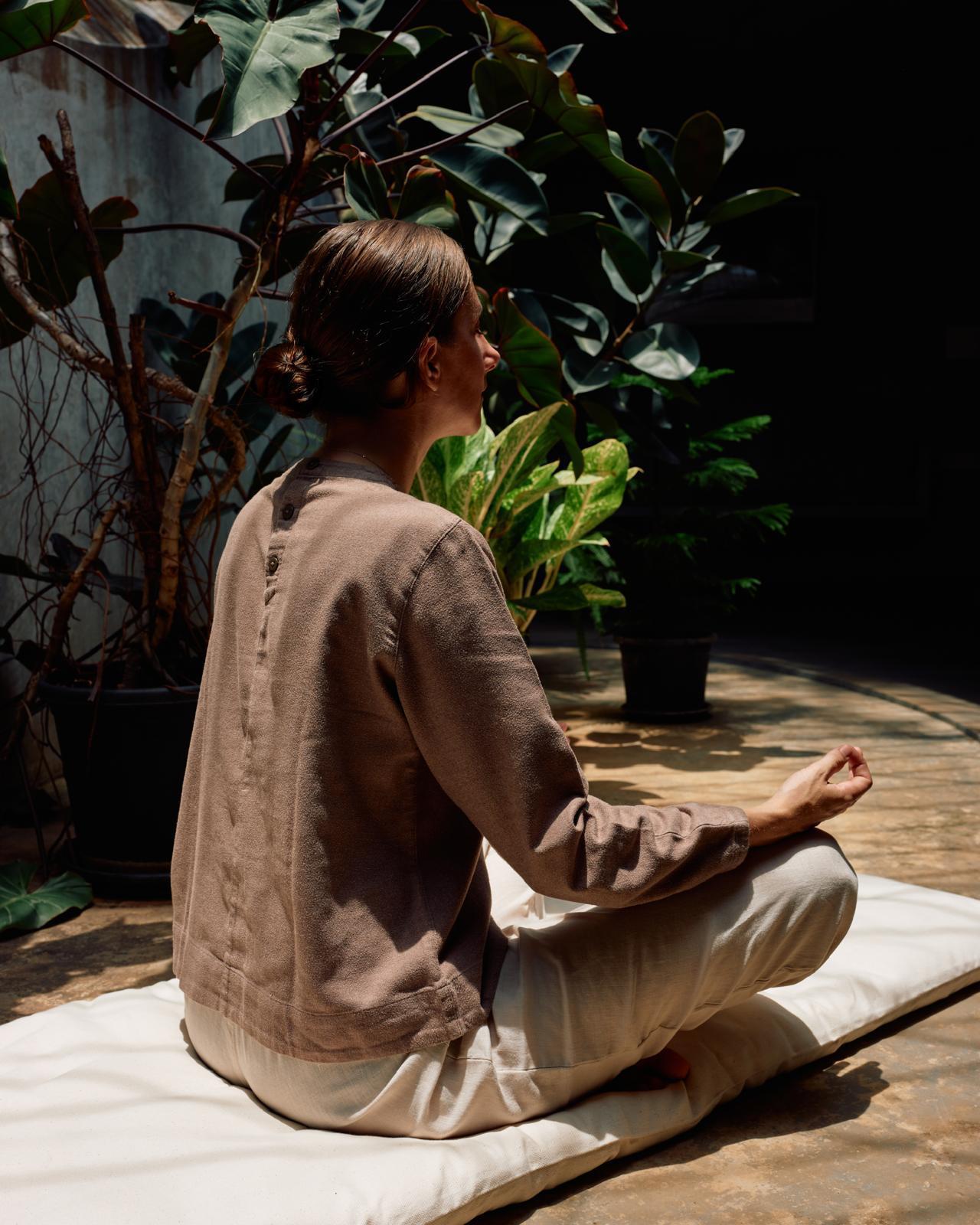 Person sitting in a meditative pose on a mat surrounded by plants