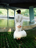 Person in a white outfit holding a large white bag in an outdoor setting with greenery.